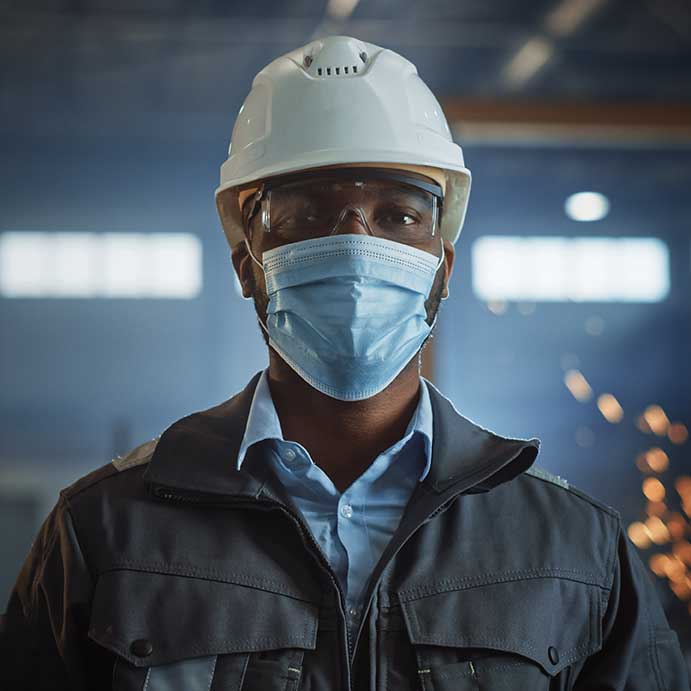 A man wearing a construction hat and a face mask while standing in a factory.