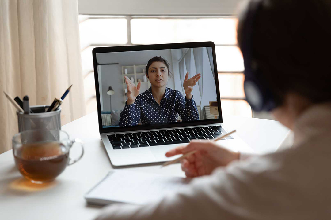 A person looking into a laptop screen as they attend a meeting, or a class and they are taking notes on a pad.