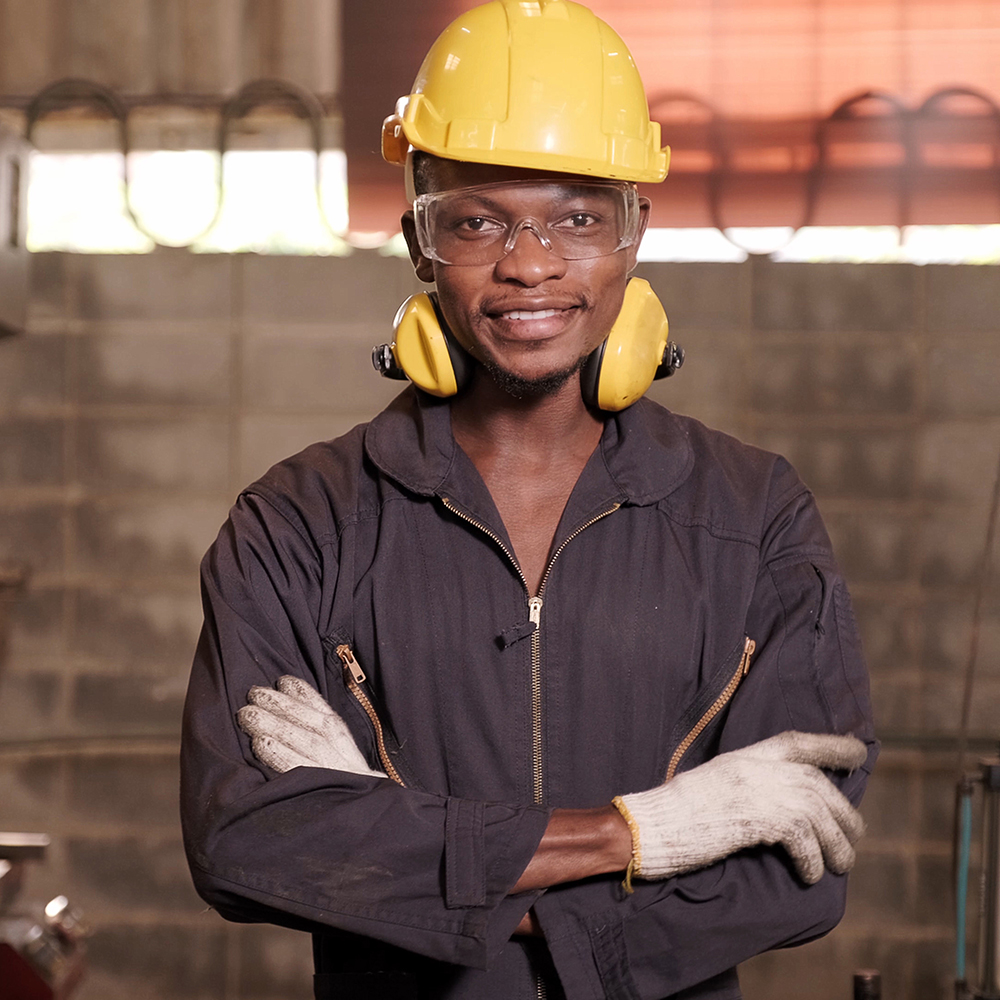 A man standing and smiling wearing a construction hat, glasses, ear guards, overalls and some gloves inside a workshop.