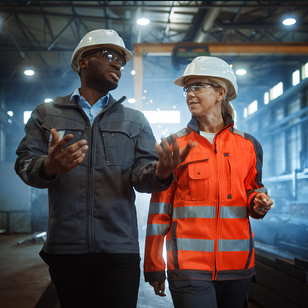 A man and a woman in protective clothing and of different races have a casual conversation at a construction site .